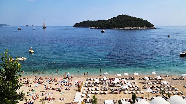 A beach with sun loungers and umbrellas, boats in the water, and a small island in the distance.