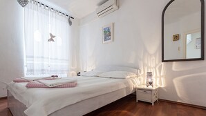 A bedroom with a large bed, a bedside table with a lamp, a window with sheer curtains, and a wall-mounted air conditioner.
