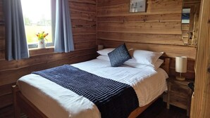 A wooden cabin bedroom with a bed, a nightstand, a lamp, a window with curtains, and a framed picture on the wall.