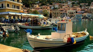 Un puerto con barcos amarrados, un edificio con terraza y una colina con viviendas al fondo.