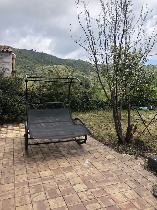 A metal outdoor bench with a mesh backrest, situated on a paved patio with a view of a green hillside and a tree with white blossoms.