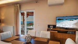 A dining area with a wooden table, white chairs, and a television displaying a beach scene. - Nereid Villa, with Heated Pool & Heated SpaWhirlpool, BBQ, Impeccable SeaViews (Rethymno)