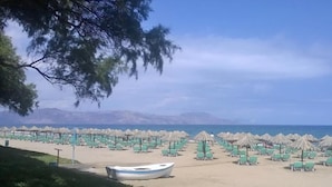 A beach with rows of green lounge chairs and thatched umbrellas, a boat on the sand, and a mountainous backdrop. - Nereid Villa, with Heated Pool & Heated SpaWhirlpool, BBQ, Impeccable SeaViews (Rethymno)