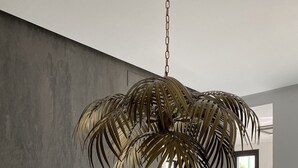 Un espace repas moderne avec une table en verre et des chaises en métal, une plante suspendue, et une vue sur la cuisine et la salle à manger.