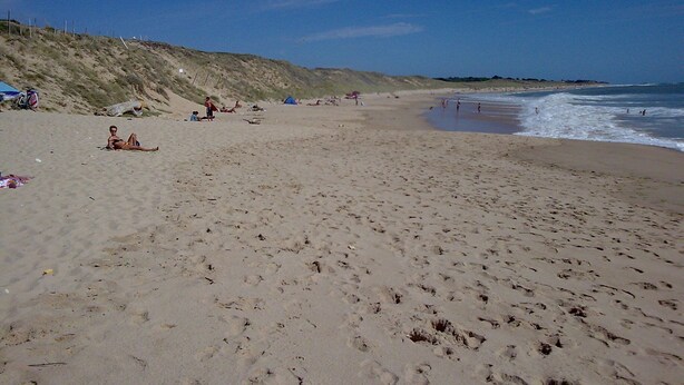 A wide sandy beach with footprints, a grassy hill, and a clear blue sky.