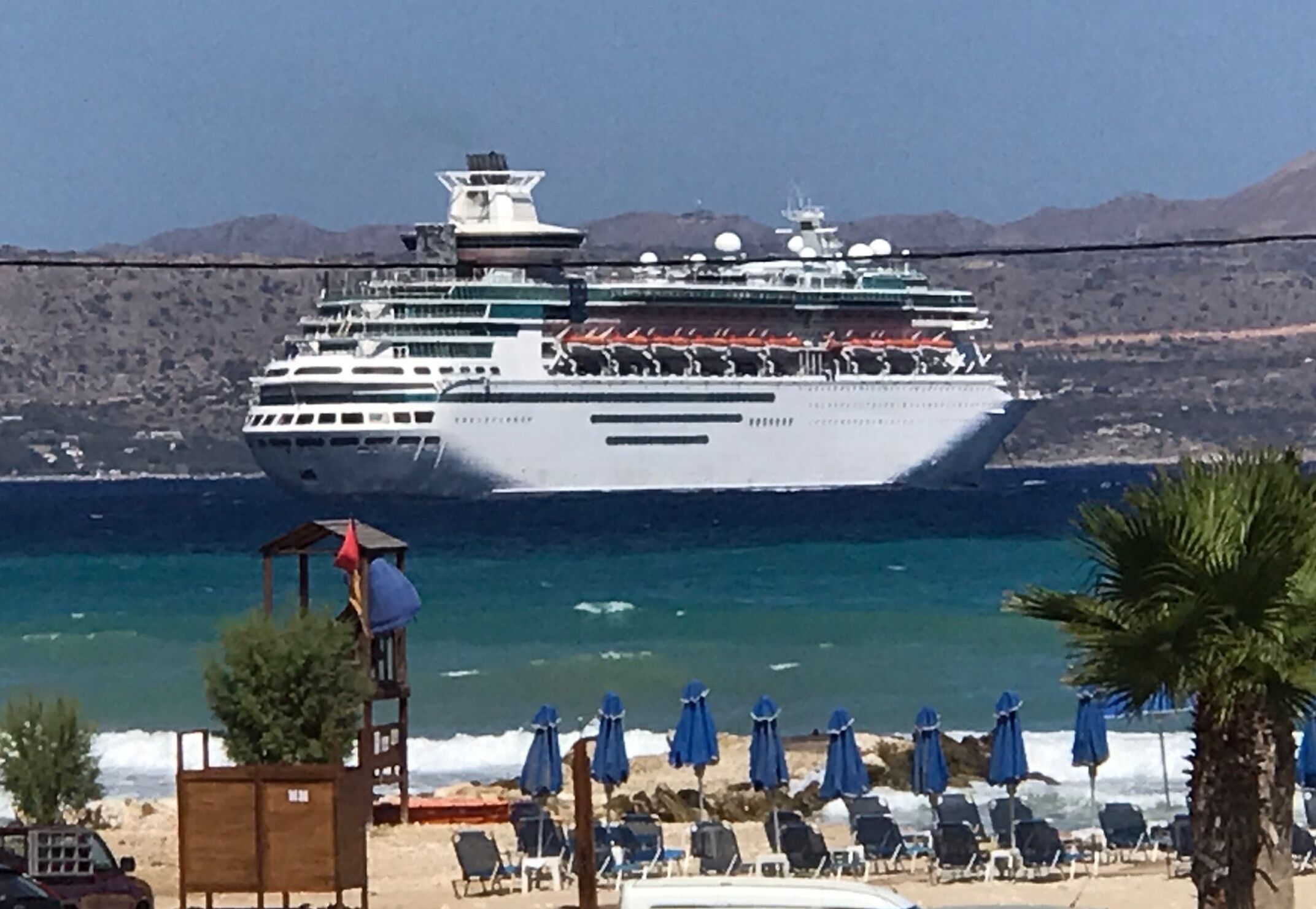 A large cruise ship docked near a beach with rows of blue umbrellas and lounge chairs.