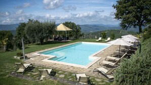 Ein rechteckiger Pool mit Steinrand, umgeben von Liegestühlen und Sonnenschirmen, vor einer Kulisse aus Bäumen und fernen Hügeln.