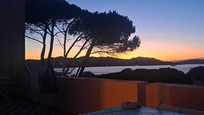 Un balcón con vista a un atardecer sobre un cuerpo de agua, árboles y colinas distantes.
