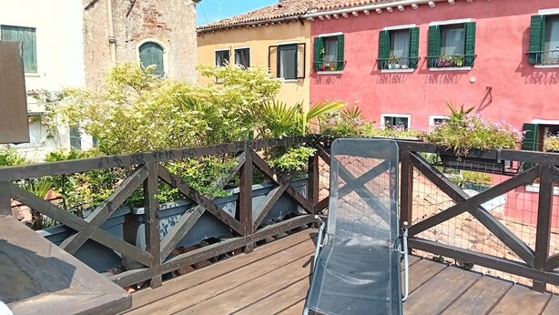 A wooden deck with a metal chair, overlooking a row of colorful buildings with green shutters and potted plants.