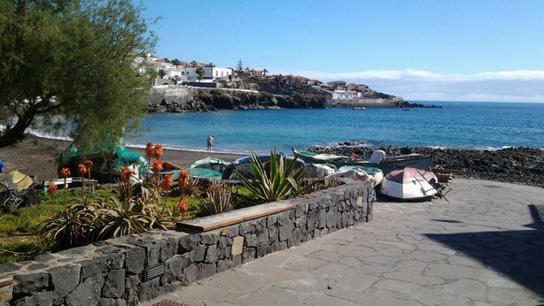 A coastal area with a stone pathway, beach umbrellas, and a view of a hillside settlement across the water. - Total Relax Apartment (Porís de Abona ( Arico ))