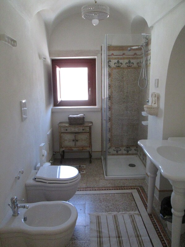 A compact bathroom with a toilet, bidet, and shower. There's a window providing natural light, and the floor has patterned tiles. - Trulli of Architect (Cisternino)