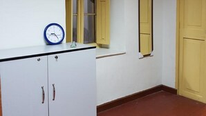 Una habitación con un armario blanco, una ventana con contraventanas amarillas, un espejo montado en la pared y un reloj.