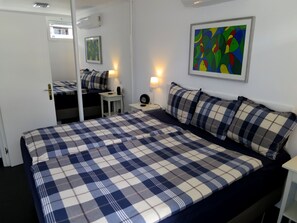A bedroom with a large bed, a bedside table with a lamp, a small table with a speaker, and a colorful abstract painting on the wall.