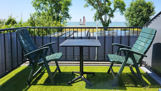A balcony with two green chairs and a small table, overlooking a beach with sailboats and trees. - Seaview & Sunset with beautiful views over the IJsselmeer (102) (Stavoren)