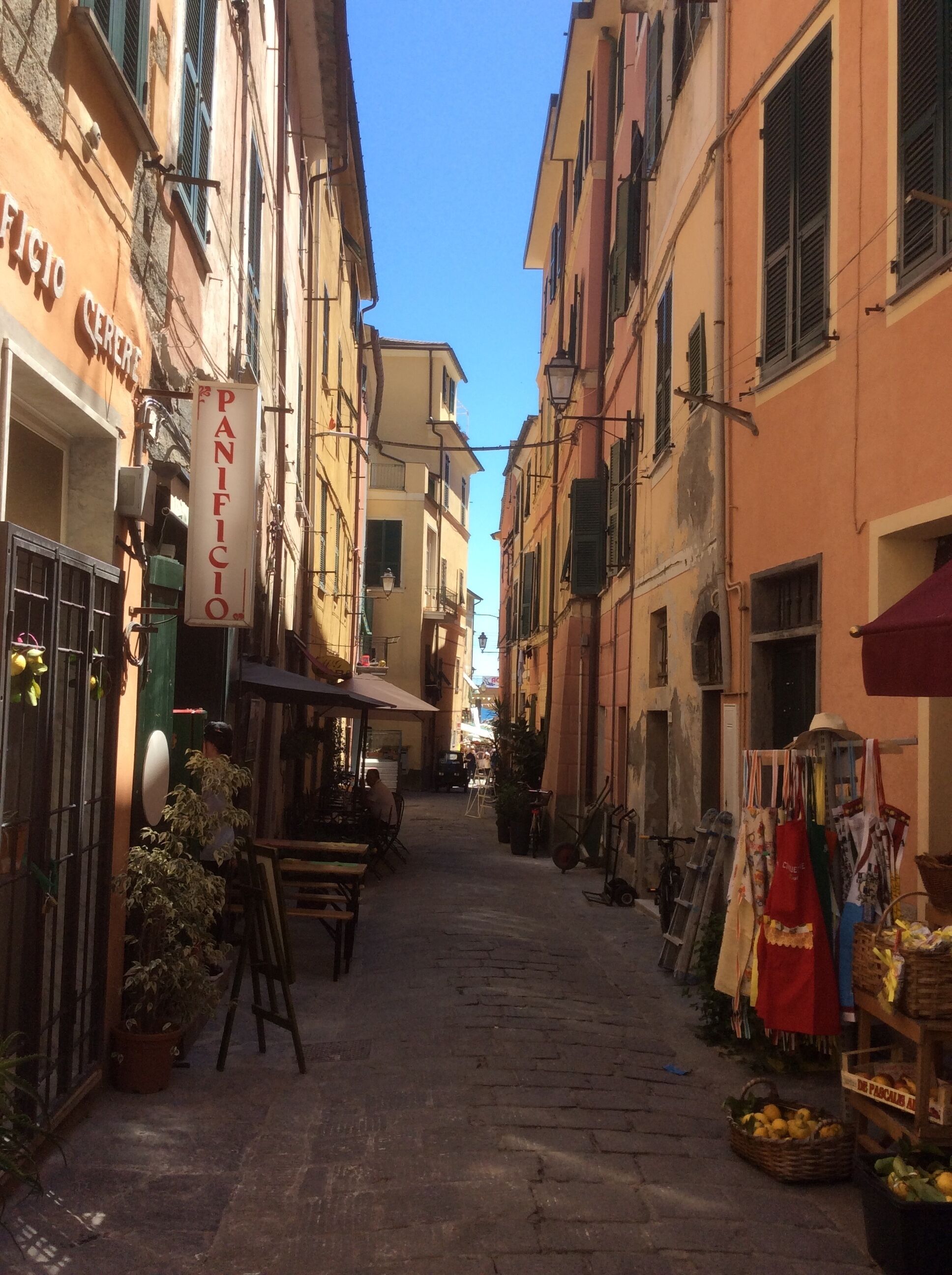 A narrow street with buildings on either side, a sign for 'PANIFICIO', and outdoor seating areas.