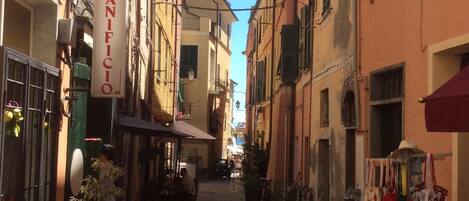 A narrow street with buildings on either side, a sign for 'PANIFICIO', and outdoor seating areas.