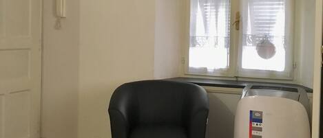 A room with a black armchair, a small ottoman, a white heater, a framed picture on the wall, and a window with curtains.