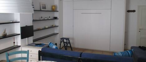 Une pièce comprenant une table à manger, un canapé bleu, des étagères ornées d’objets décoratifs, une armoire et un téléviseur.