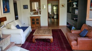 A living room with a white sofa, a brown leather sofa, a wooden coffee table, and a red patterned rug. There is a wooden cabinet with a mirror, a dining area with chairs, and a kitchen visible through an open doorway. - Nice house with view (Vero)