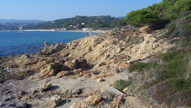 Un paisaje costero rocoso con un mar azul cristalino, un pequeño velero y un sendero que lleva a una colina con casas al fondo.