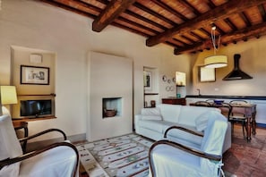 A living room with a white sofa, wooden chairs, a television, a fireplace, and a kitchen area with a hanging light fixture. - Country House Nazzano in Vineyards/ near San Gimignano Florence Siena - Ro (Gambassi Terme)