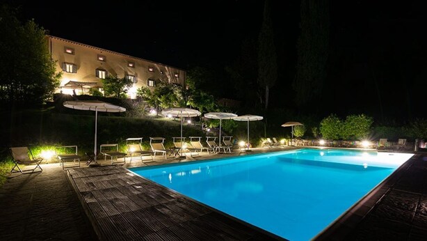 A nighttime swimming pool with lounge chairs and umbrellas, surrounded by a wooden deck and a building with lit windows.