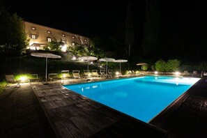 A nighttime swimming pool with lounge chairs and umbrellas, surrounded by a wooden deck and a building with lit windows. - Villa Catarsena with private pool in Tuscany (Bibbiena)
