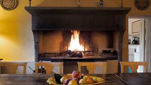 A fireplace with a fire, a table with a fruit bowl, and a kitchen area in the background.