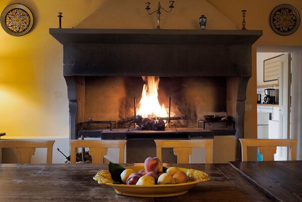 A fireplace with a fire, a table with a fruit bowl, and a kitchen area in the background. - Villa Catarsena with private pool in Tuscany (Bibbiena)