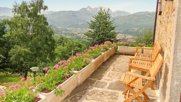 A stone terrace with a view of mountains, a wooden bench, and flower beds.