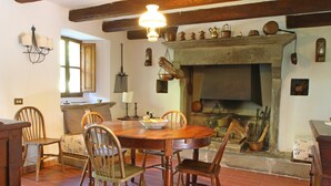 A rustic dining area with a wooden table and chairs, a fireplace, and traditional kitchen utensils displayed on the wall.