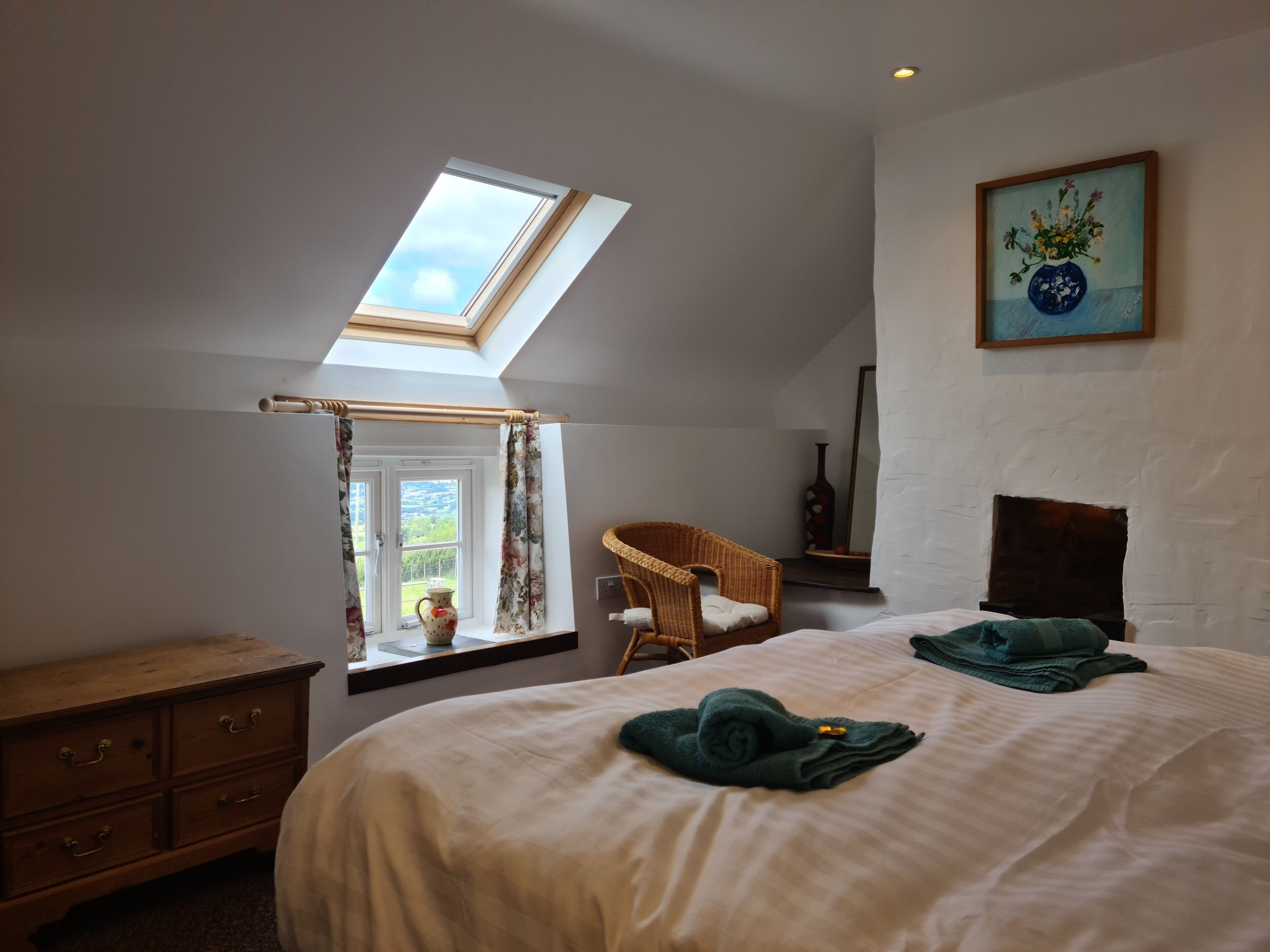 Une chambre avec une lucarne, un lit avec des serviettes vertes, une chaise en osier et un tableau au mur.