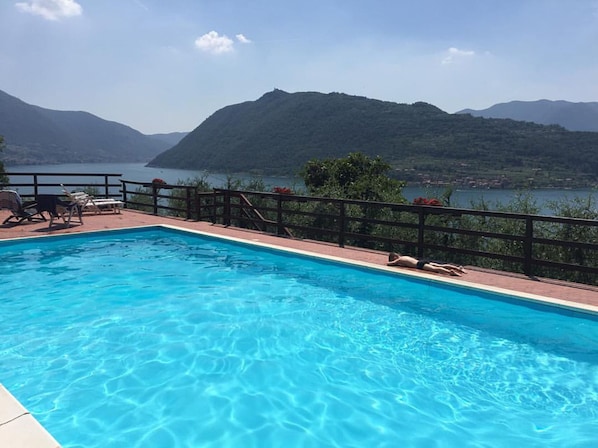 Una piscina con acqua cristallina, circondata da un pontile in legno e una ringhiera. Sullo sfondo, montagne e un corpo d'acqua.