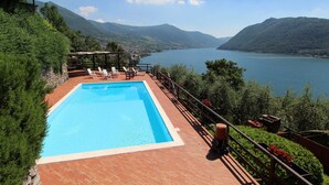 A rectangular pool with a wooden deck, surrounded by greenery and overlooking a lake with mountains in the background. - Two-room apartment in Residence with swimming pool and panoramic lake view in Sale Marasino (Sale Marasino)