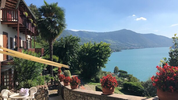 A balcony with a stone wall, potted plants, and a view of a lake and mountains. - Two-room apartment in Residence with swimming pool and panoramic lake view in Sale Marasino (Sale Marasino)