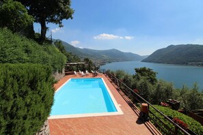 Una piscina rettangolare con bordo in legno e ringhiera, circondata da vegetazione rigogliosa e affacciata su un lago con montagne sullo sfondo.