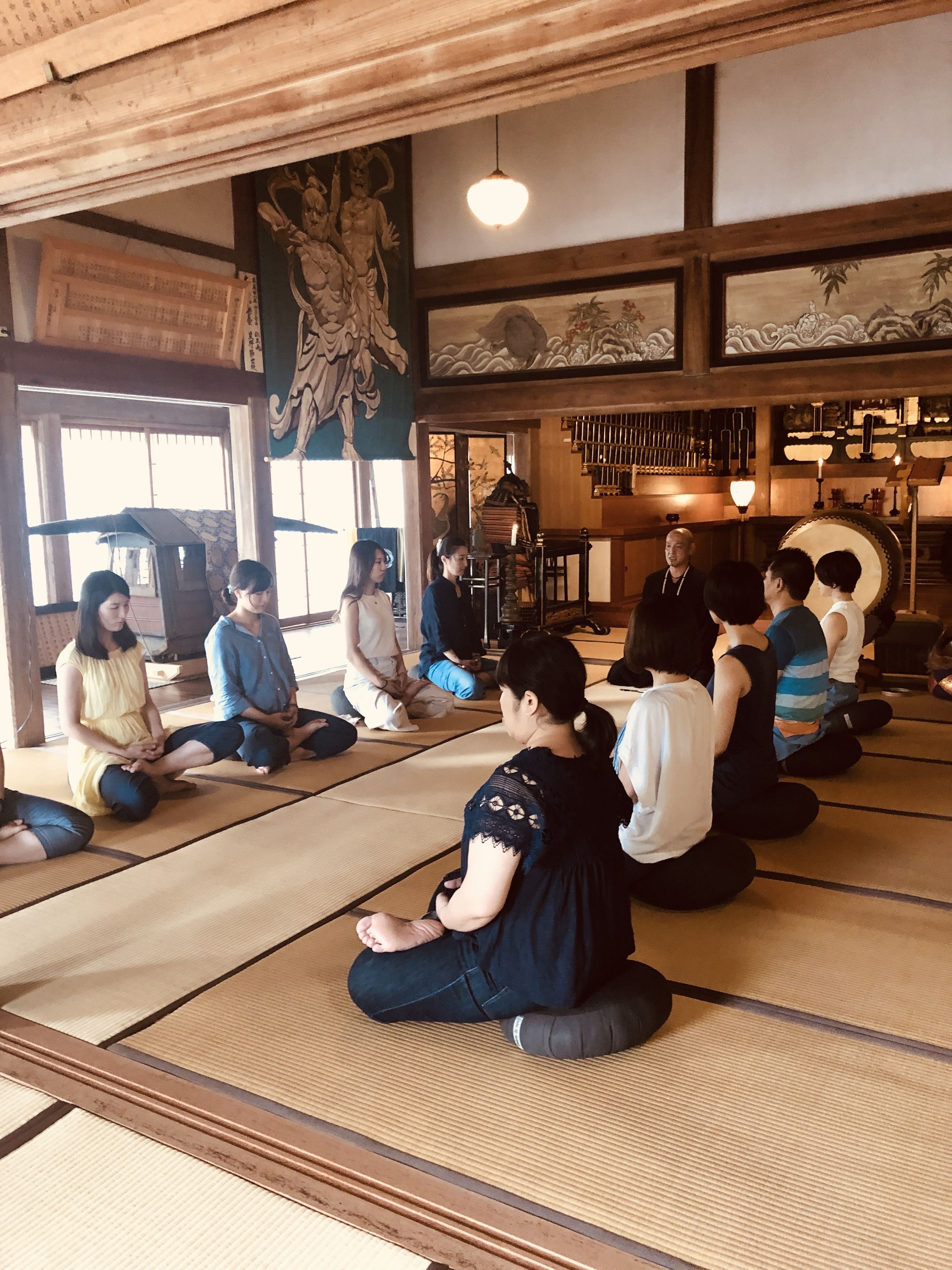 A tatami mat room with a group of people seated in a circle, a traditional Japanese room with tatami flooring, a hanging light fixture, and a mural on the wall.