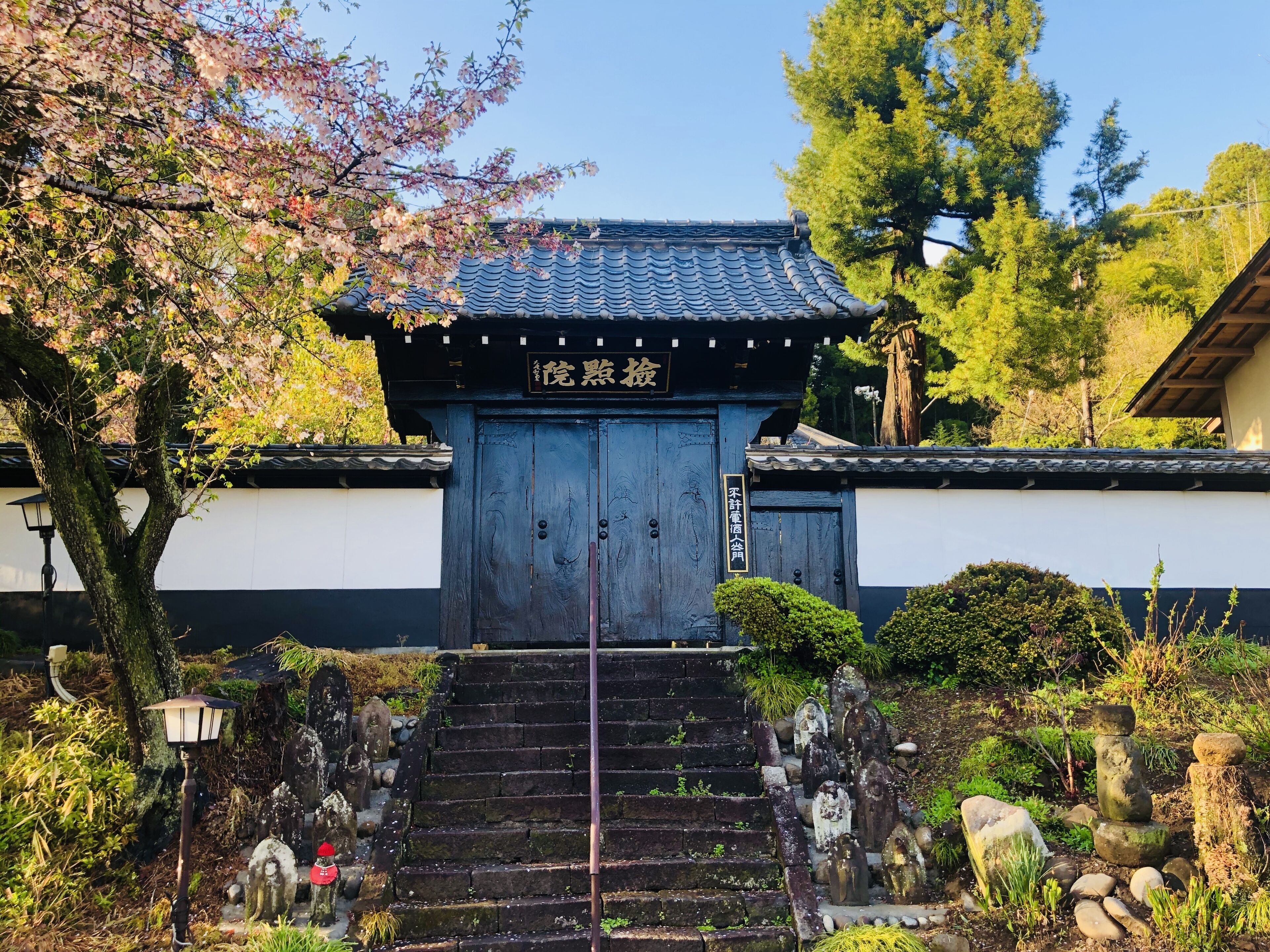 Um portão tradicional japonês com uma escadaria de madeira que leva até ele, cercado por lampiões de pedra e um jardim bem cuidado.