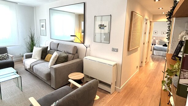 Ein modernes Wohnzimmer mit grauem Sofa, einem kleinen runden Tisch und einem Glastisch. An der Wand befindet sich ein großer Spiegel und rechts eine Pflanze.