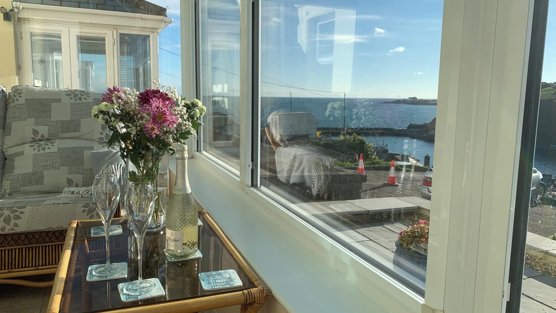 Un balcone con vista sul mare, una sedia in vimini, un tavolino con un vaso di fiori e una bottiglia di champagne.