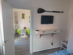 A room with a bed, a mounted TV, a wall-mounted paddle, a desk with drawers, and a dining area visible through an open door.
