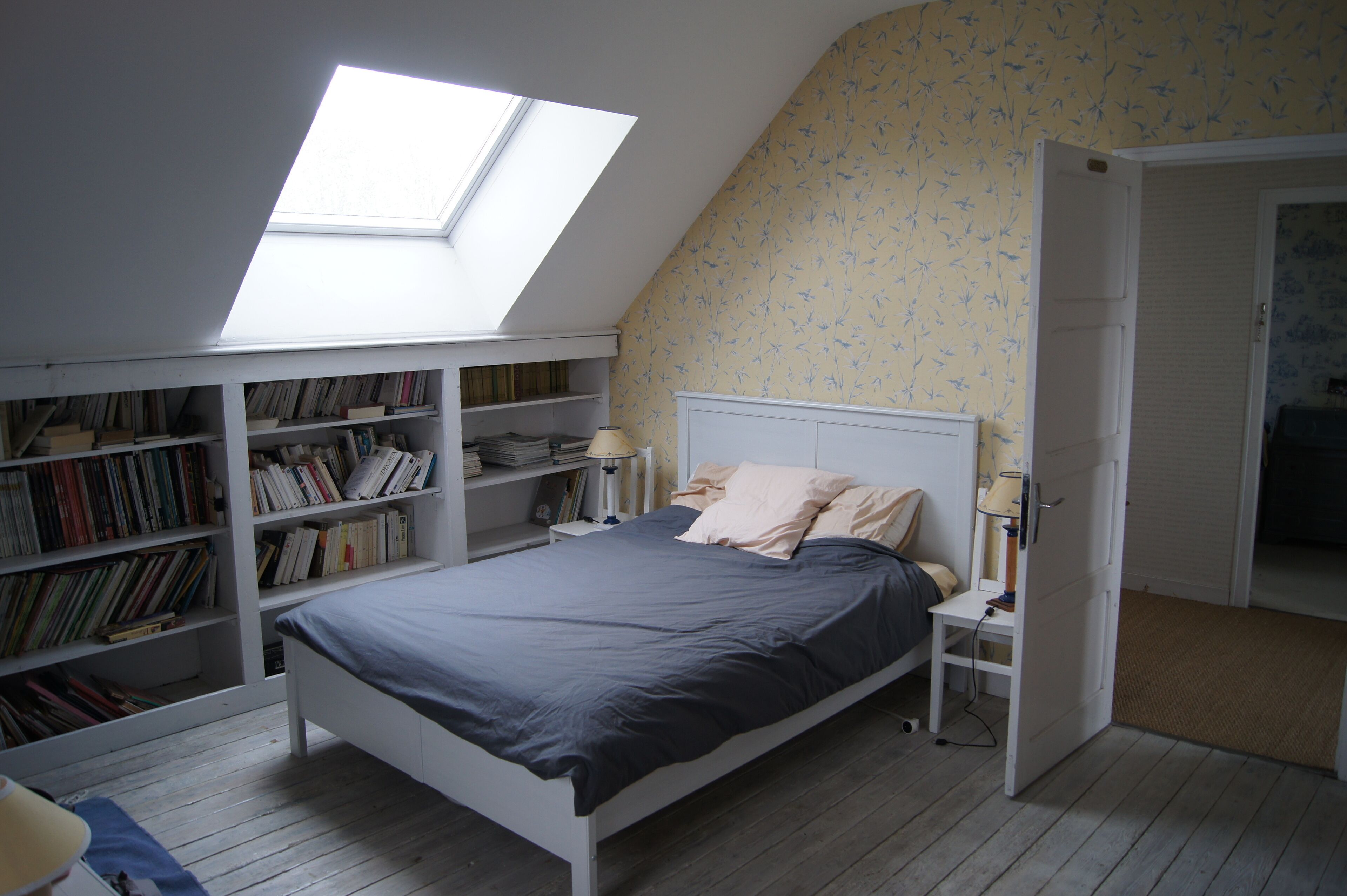 Een slaapkamer met dakraam, een bed, boekenkasten en een deur naar een andere kamer.