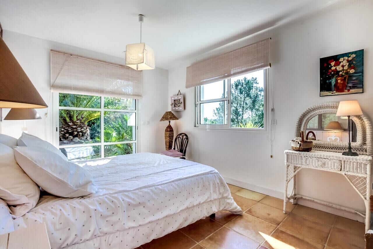 Une chambre à coucher avec un lit, deux fenêtres avec des rideaux, une table de chevet avec une lampe, un fauteuil en osier et un tableau accroché au mur.