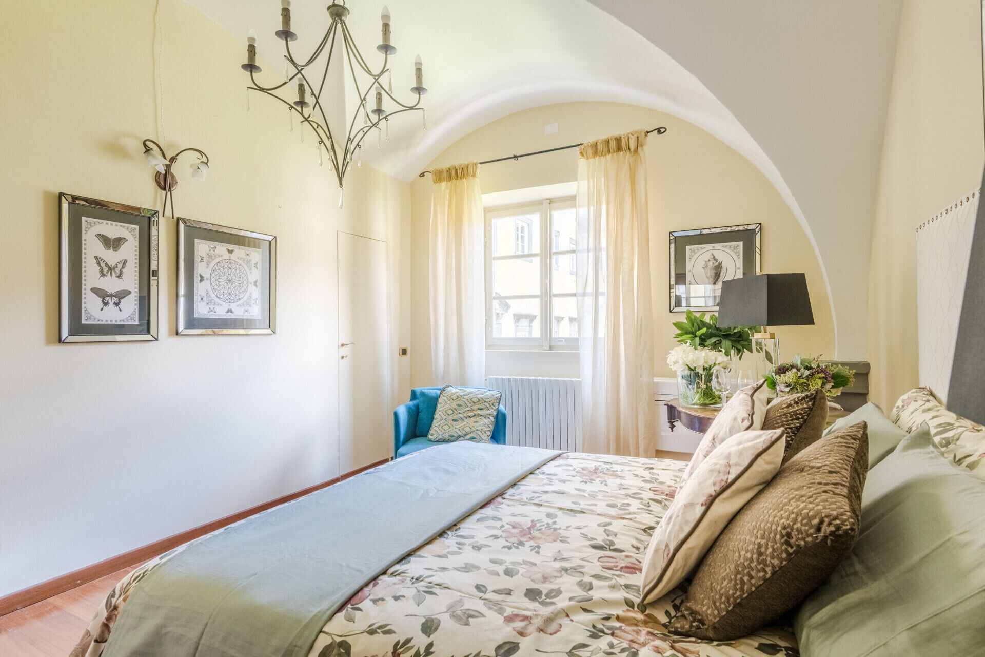 Een slaapkamer met een groot bed, een kroonluchter, lijstwerken, een raam met gordijnen en een nachtkastje met een lamp en bloemen.