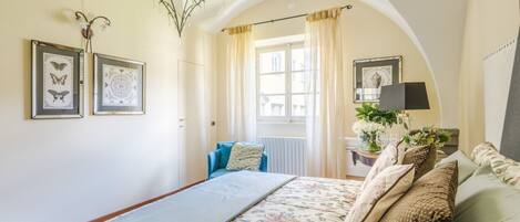 Een slaapkamer met een groot bed, een kroonluchter, lijstwerken, een raam met gordijnen en een nachtkastje met een lamp en bloemen.