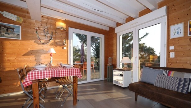 Interior em madeira com uma mesa de jantar posta para quatro pessoas, um sofá e uma porta de vidro que dá para o exterior.