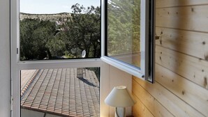 Quarto com uma grande janela, uma cama, uma mesa de cabeceira com abajur, e vista para um telhado e árvores lá fora.