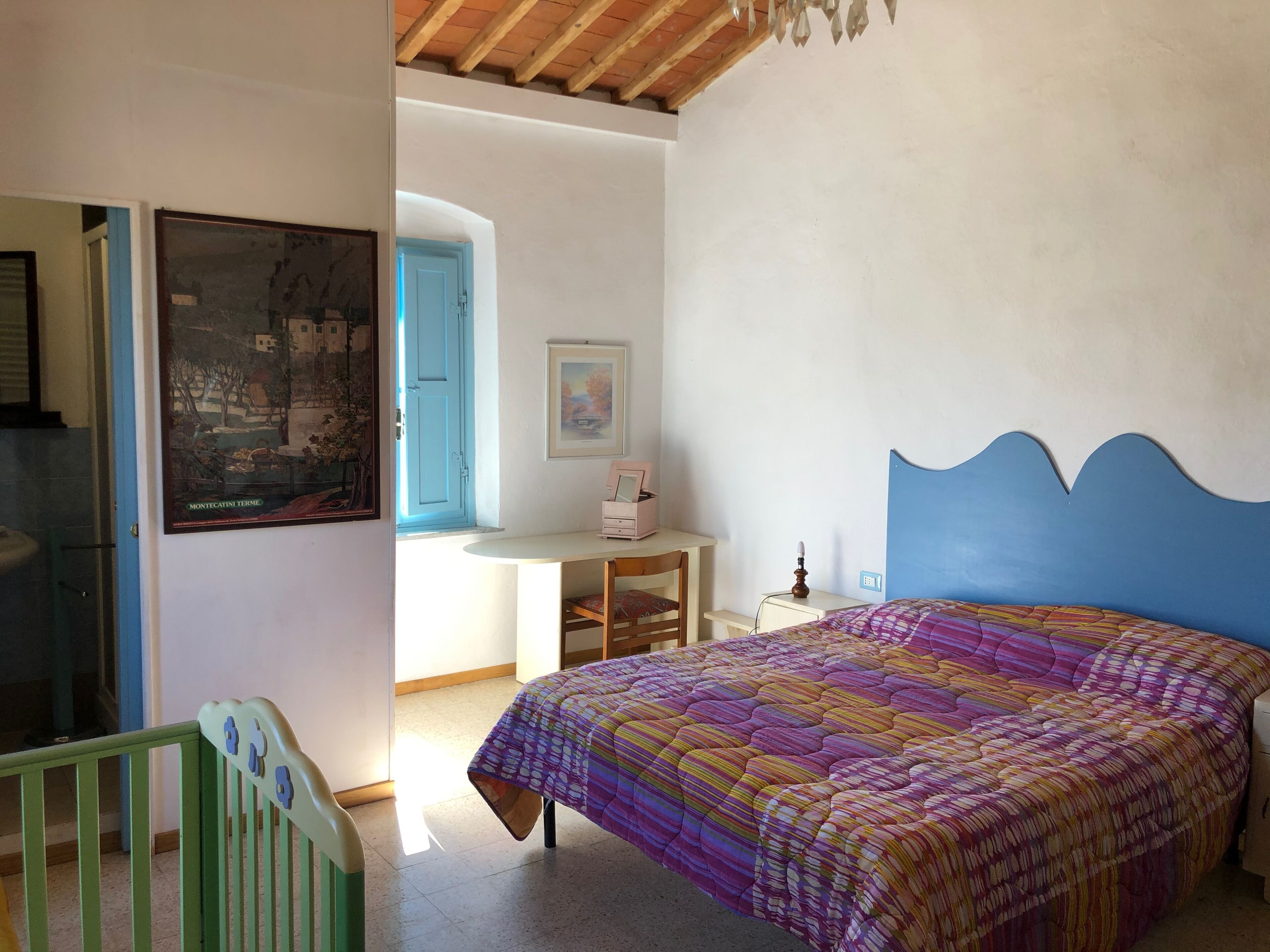A bedroom with a colorful bedspread, a blue headboard, a desk with a chair, and a framed picture on the wall.