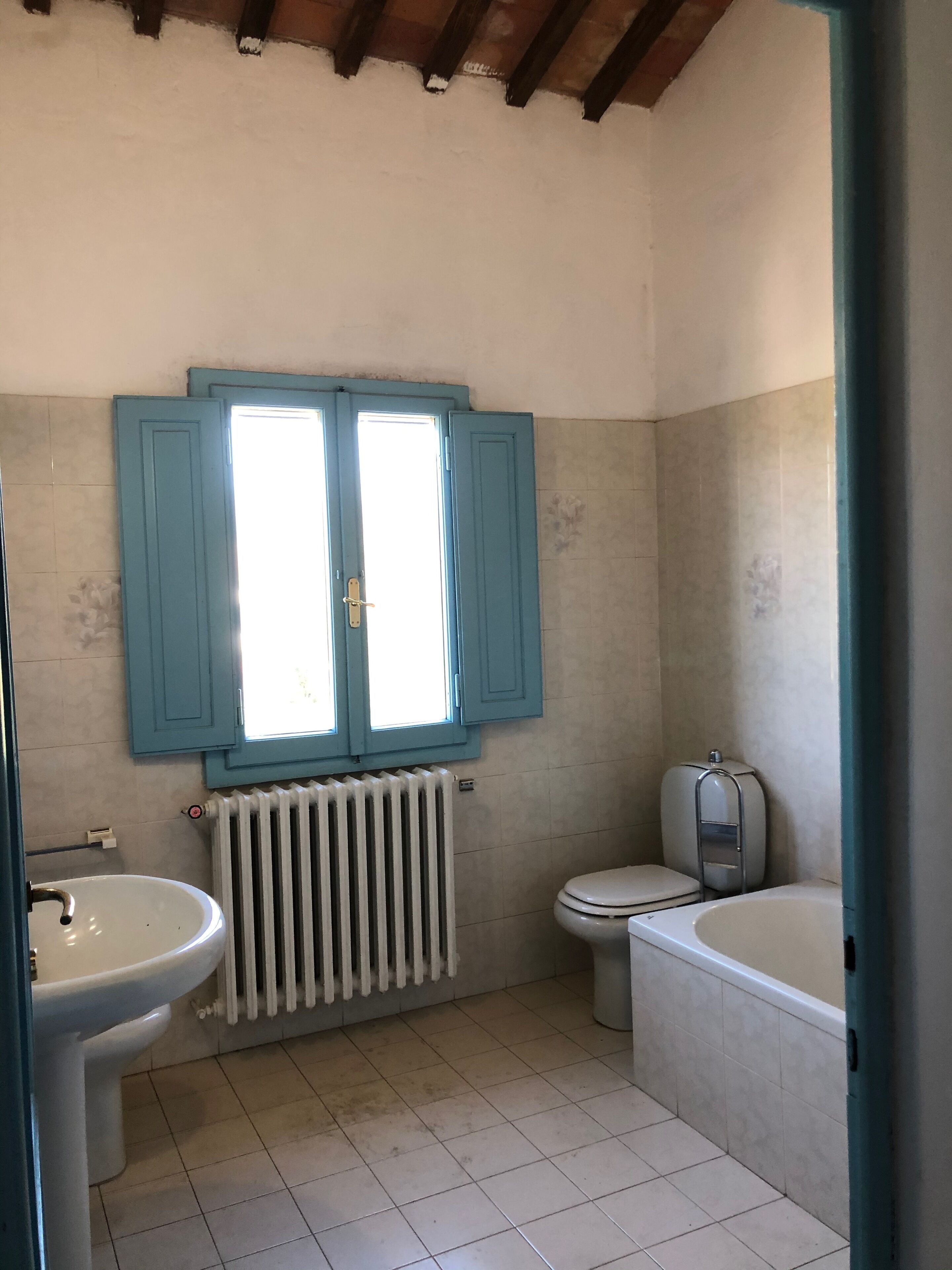 A bathroom with a white sink, toilet, and bathtub, a window with blue shutters, and a radiator.