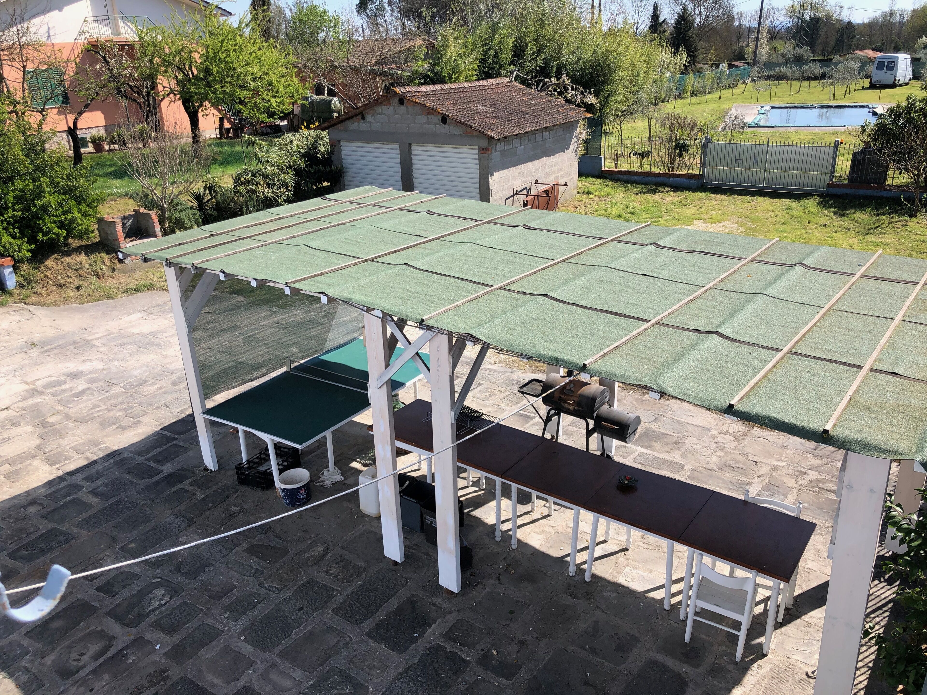 Ein überdachter Außenbereich mit Tisch und Bänken, umgeben von einem Garten und einem kleinen Gebäude.